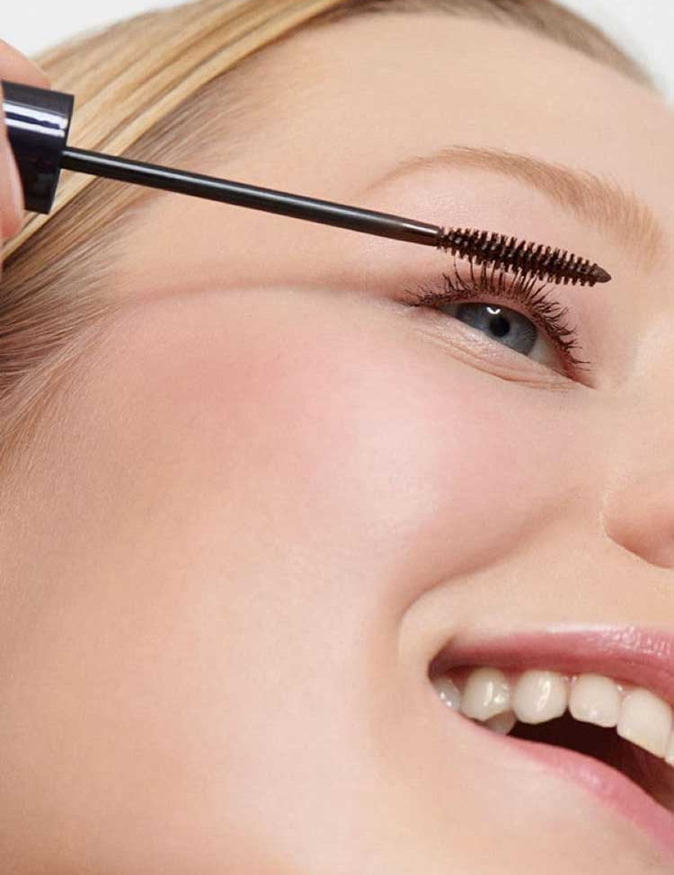 A close-up of a smiling person with blue eyes applying Dr Hauschka Defining Mascara 6ml—suitable for sensitive eyes—to their upper lashes. Their skin is smooth, and the mascara wand is held gently near their eyelashes.