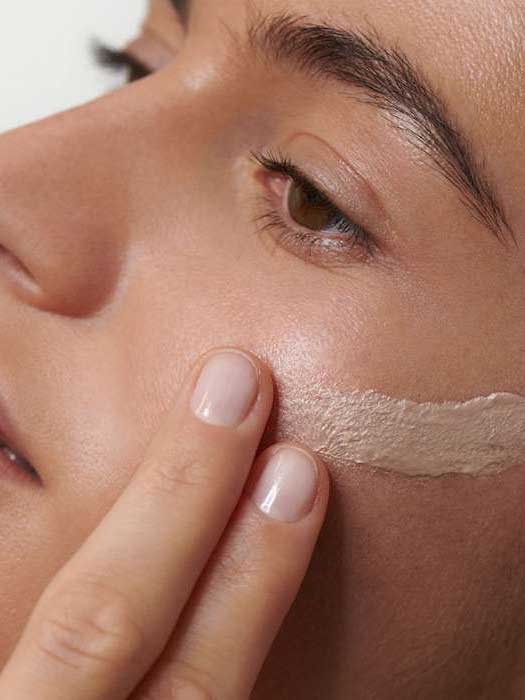 A close-up of someone applying Dr Hauschka Illuminating Fluid 30ml to their cheek with their fingers, highlighting the gentle application and natural glow for radiant, healthy-looking skin.