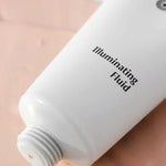 A white tube of Dr Hauschka Illuminating Fluid 30ml rests on a peach surface, cap off, with the hydrating highlighter gently dispensed for a natural glow.