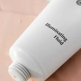 A white tube of Dr Hauschka Illuminating Fluid 30ml rests on a peach surface, cap off, with the hydrating highlighter gently dispensed for a natural glow.