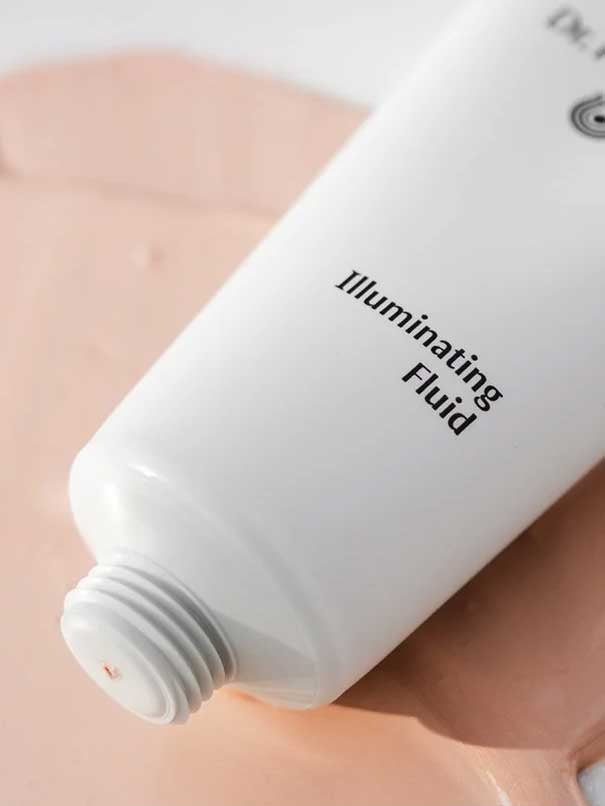 A white tube of Dr Hauschka Illuminating Fluid 30ml rests on a peach surface, cap off, with the hydrating highlighter gently dispensed for a natural glow.