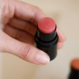 A hand holds Dr Hauschka Lip to Cheek Tint & Highlighter - Gold 6.1g with the cap off, revealing its natural multi-purpose tint; another similar stick is softly blurred in the background.