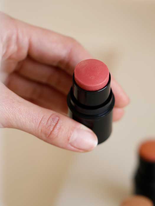 A hand holds Dr Hauschka Lip to Cheek Tint & Highlighter - Gold 6.1g with the cap off, revealing its natural multi-purpose tint; another similar stick is softly blurred in the background.