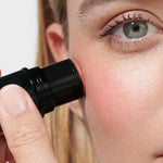 A person applies Dr Hauschka Lip to Cheek Tint - Apricot 6.1g to their cheek with a makeup stick. The close-up image highlights one eye, the cheek, and the hand holding the tint stick.