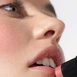 A close-up shows a person smoothing Dr Hauschka Lip to Cheek Tint - Apricot 6.1g onto their lips, with smooth skin and an upward gaze, highlighting the product’s natural and versatile use for both lips and cheeks.