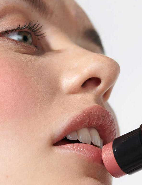 A close-up shows a person smoothing Dr Hauschka Lip to Cheek Tint - Apricot 6.1g onto their lips, with smooth skin and an upward gaze, highlighting the product’s natural and versatile use for both lips and cheeks.
