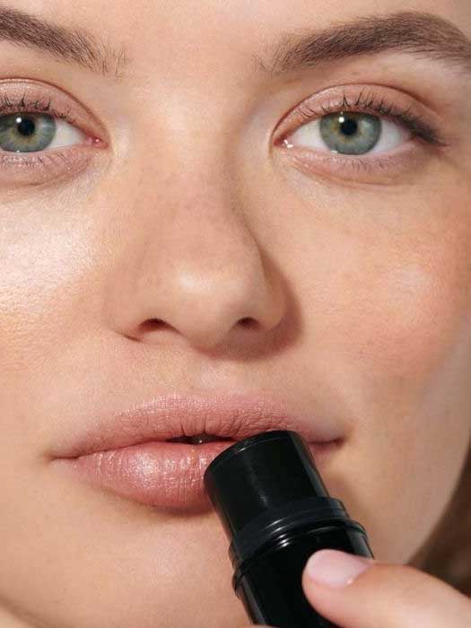 A close-up of a person applying Dr Hauschka Lip to Cheek Tint & Highlighter - Gold 6.1g to their lips, with clear skin and green eyes, showcases its gold shimmer and versatile use for both lips and cheeks.