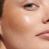 Close-up of a woman's face with smooth, glowing skin, natural makeup, and Dr Hauschka Lip to Cheek Tint & Highlighter - Gold 6.1g on her cheekbone; only one eye and part of her lips are visible.
