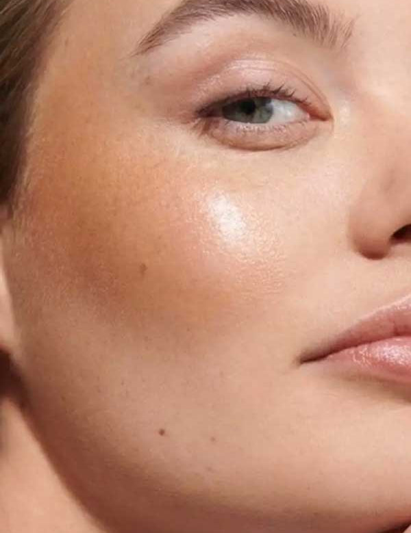 Close-up of a woman's face with smooth, glowing skin, natural makeup, and Dr Hauschka Lip to Cheek Tint & Highlighter - Gold 6.1g on her cheekbone; only one eye and part of her lips are visible.