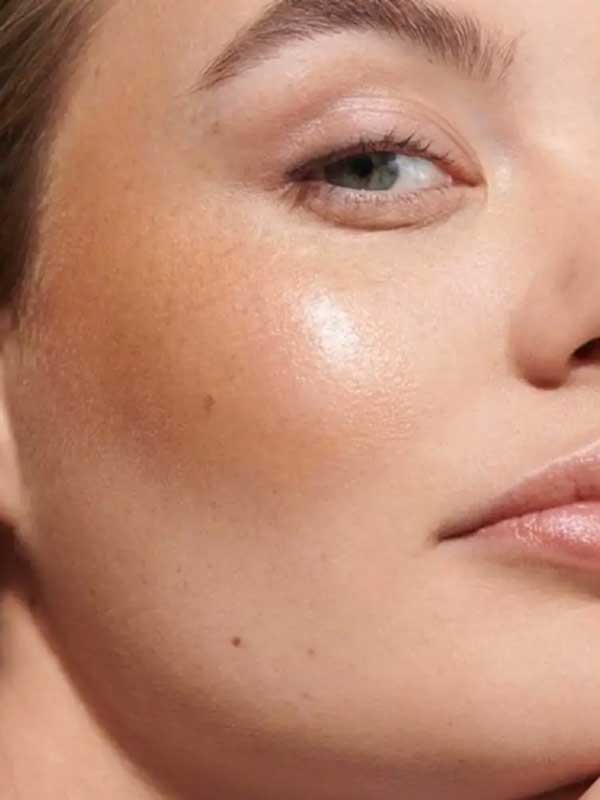 Close-up of a woman's face with smooth, glowing skin, natural makeup, and Dr Hauschka Lip to Cheek Tint & Highlighter - Gold 6.1g on her cheekbone; only one eye and part of her lips are visible.