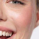 Close-up of a smiling person with clear skin, greenish eyes, and light makeup. White teeth and healthy skin glow, accented by Dr Hauschka Lip to Cheek Tint - Rosewood 6.1g for a natural, joyful look on lips and cheeks.