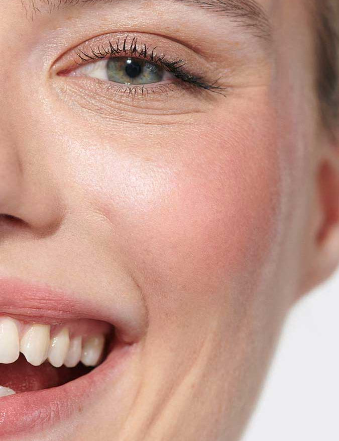 Close-up of a smiling person with clear skin, greenish eyes, and light makeup. White teeth and healthy skin glow, accented by Dr Hauschka Lip to Cheek Tint - Rosewood 6.1g for a natural, joyful look on lips and cheeks.