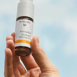 A hand holds up a bottle of Dr Hauschka Translucent Bronzing Tint 18ml, featuring natural mineral pigments, against a blue sky with soft clouds.