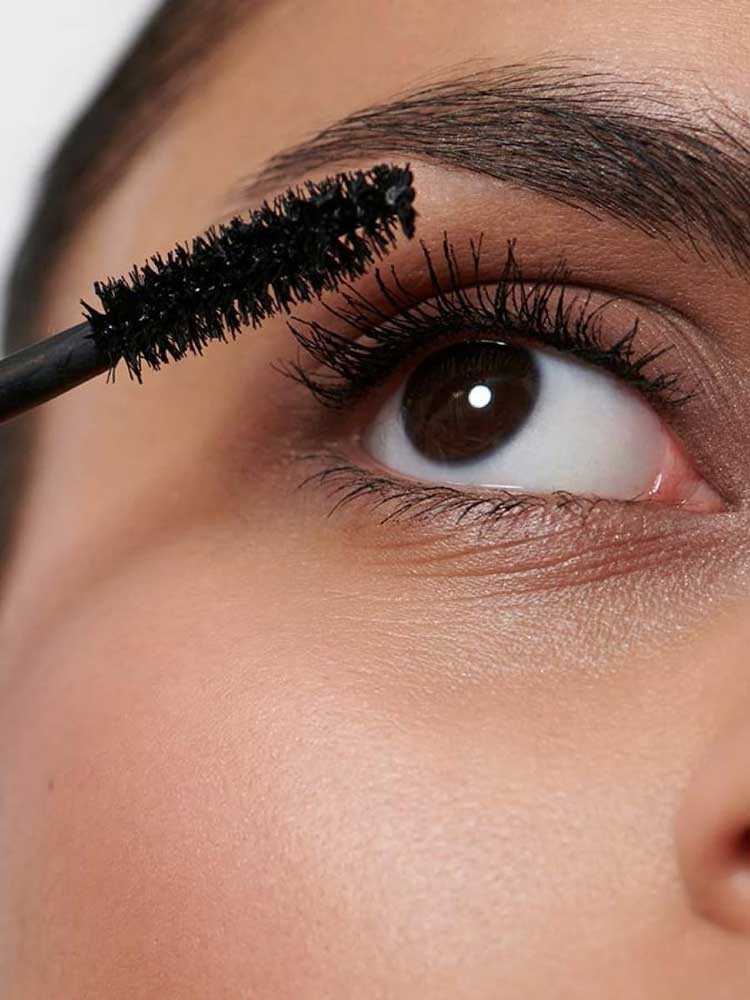 A close-up shows someone applying Dr Hauschka Volume Mascara 8ml to their upper lashes with a wand. Their eye is open, and their smooth, illuminated skin is visible.