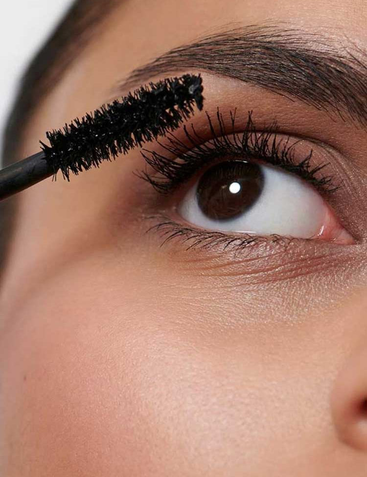 A close-up shows someone applying Dr Hauschka Volume Mascara 8ml to their upper lashes with a wand. Their eye is open, and their smooth, illuminated skin is visible.