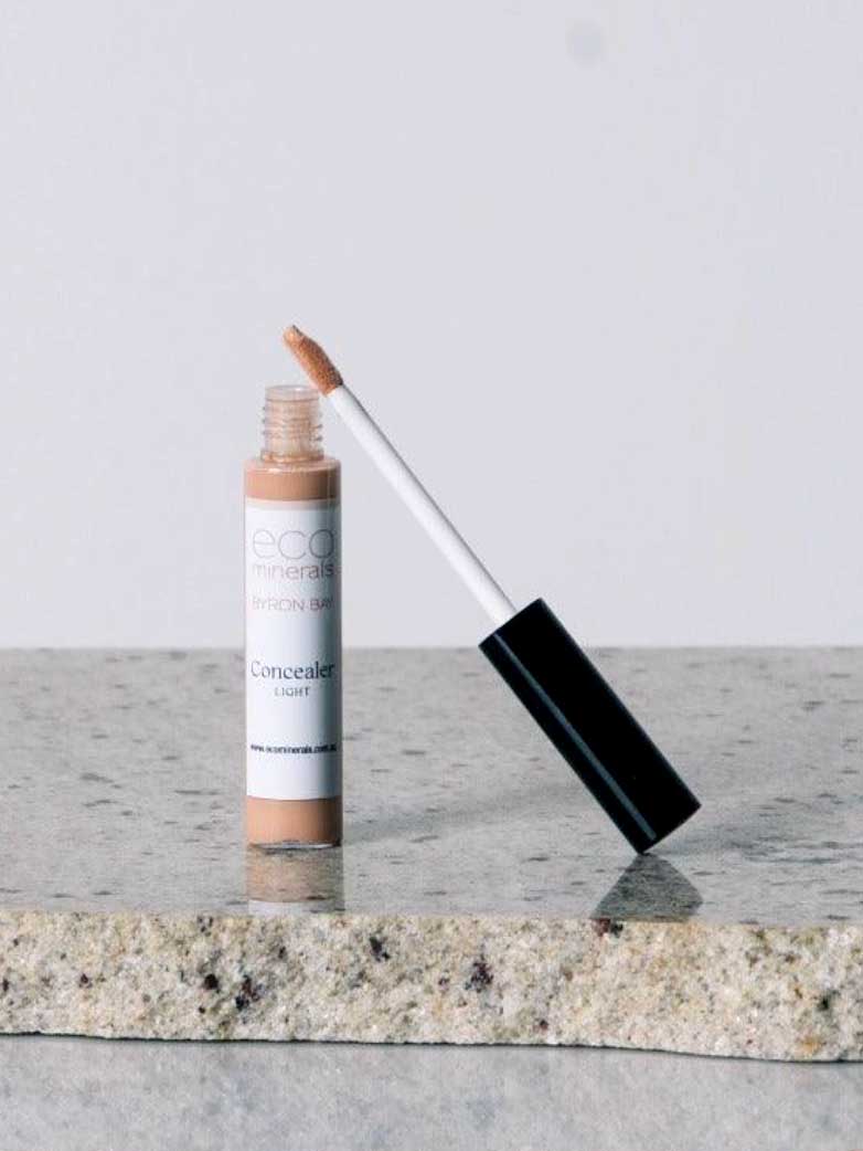 The Eco Minerals Mineral Concealer 5g, featuring a black cap and applicator wand, stands upright on a speckled stone surface with a plain light gray background.