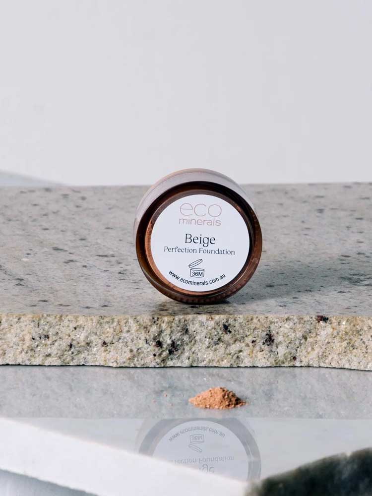 A small round container of Eco Minerals Perfection Dewy Mineral Foundation 5g sits on granite, with some loose powder spilled in front against a plain, neutral background.