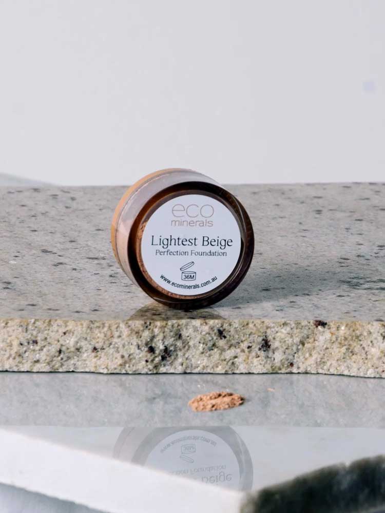 A small round container labeled Eco Minerals Perfection Dewy Mineral Foundation 5g sits on a stone surface with some loose powder in front, offering a natural dewy finish.