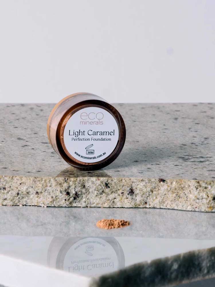 A small round container labeled “Eco Minerals Perfection Dewy Mineral Foundation 5g” rests on a stone surface with a mound of light brown powder in front, offering a dewy finish and enhancing natural radiance.