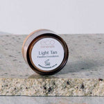 A small round container of Eco Minerals Flawless Matte Mineral Foundation 5g rests on a stone surface, with some loose powder scattered beside it.