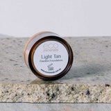 A small round container of Eco Minerals Flawless Matte Mineral Foundation 5g rests on a stone surface, with some loose powder scattered beside it.