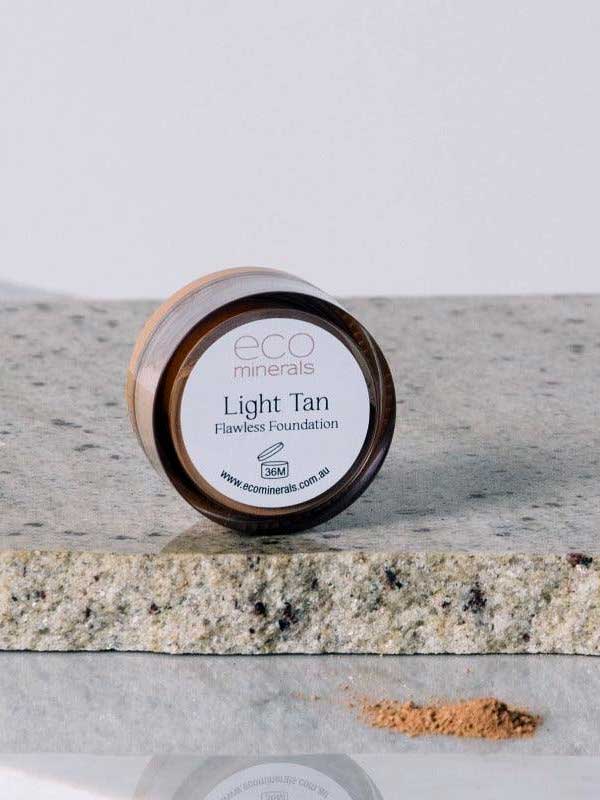 A small round container of Eco Minerals Flawless Matte Mineral Foundation 5g rests on a stone surface, with some loose powder scattered beside it.