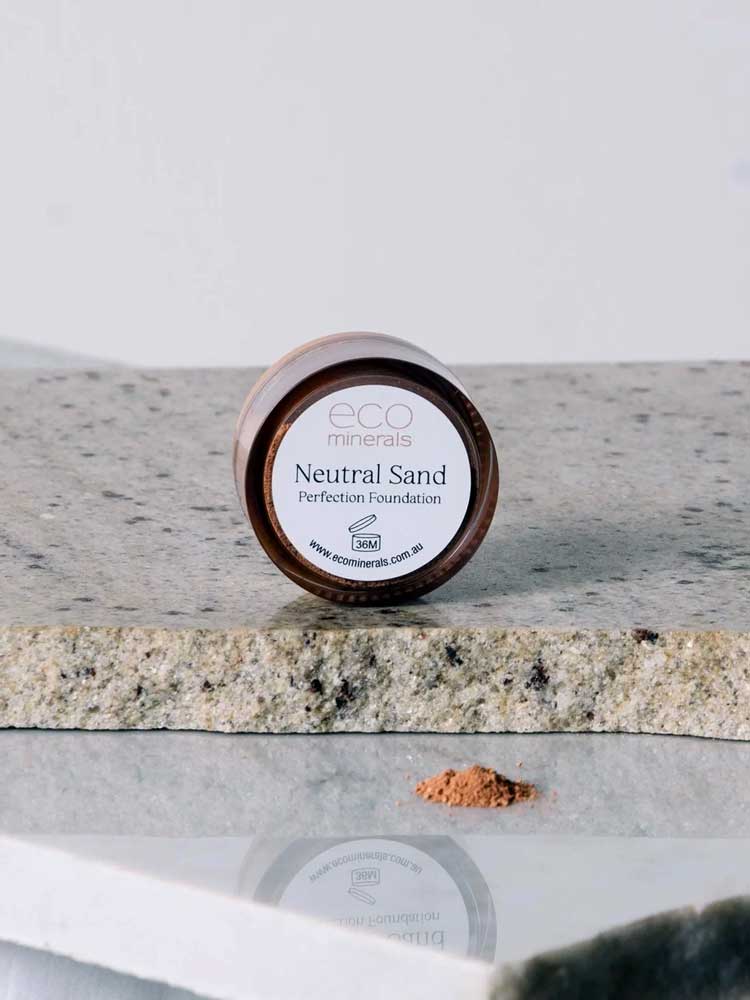 A small jar labeled Eco Minerals Perfection Dewy Mineral Foundation 5g sits on a gray stone surface, with a small pile of loose powder in front of it.