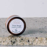 A 5g jar of Eco Minerals Flawless Matte Mineral Foundation rests on a speckled stone, some loose powder nearby. It has a brown lid, white label, delivers a matte finish, and offers natural SPF25 protection.