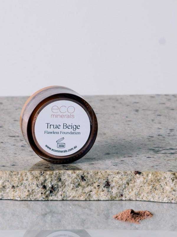 A 5g jar of Eco Minerals Flawless Matte Mineral Foundation rests on a speckled stone, some loose powder nearby. It has a brown lid, white label, delivers a matte finish, and offers natural SPF25 protection.