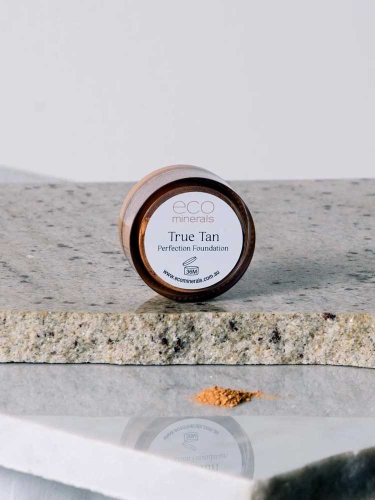 Eco Minerals Perfection Dewy Mineral Foundation 5g, in a round container on stone, is shown with loose powder in front—ideal for a dewy finish and natural SPF25 protection.