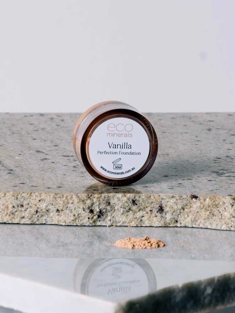 A small round container of Eco Minerals Perfection Dewy Mineral Foundation 5g rests on a granite surface, with some loose powder foundation spilled in front.