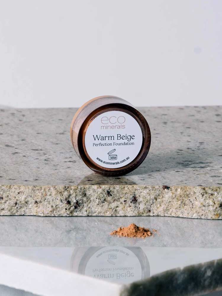 A 5g container of Eco Minerals Perfection Dewy Mineral Foundation rests on a stone surface, with some loose powder spilled beside it.