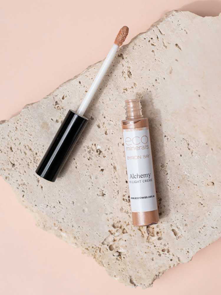 An open tube of Eco Minerals Mineral Alchemy High Light Creme 8g, a vegan highlighter with a wand applicator, rests on a textured stone slab against a soft peach backdrop for a luminous glow.