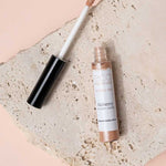 An open tube of Eco Minerals Mineral Alchemy High Light Creme 8g, a vegan highlighter with a wand applicator, rests on a textured stone slab against a soft peach backdrop for a luminous glow.