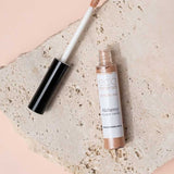 An open tube of Eco Minerals Mineral Alchemy High Light Creme 8g, a vegan highlighter with a wand applicator, rests on a textured stone slab against a soft peach backdrop for a luminous glow.