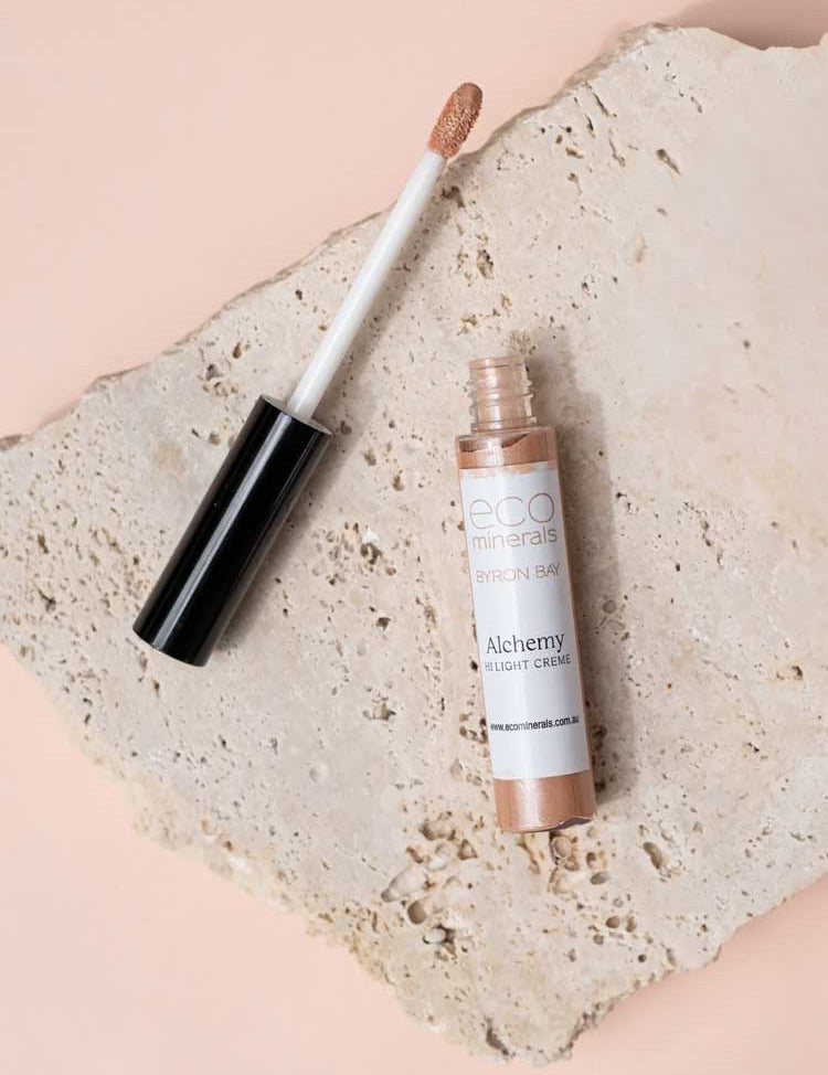 An open tube of Eco Minerals Mineral Alchemy High Light Creme 8g, a vegan highlighter with a wand applicator, rests on a textured stone slab against a soft peach backdrop for a luminous glow.