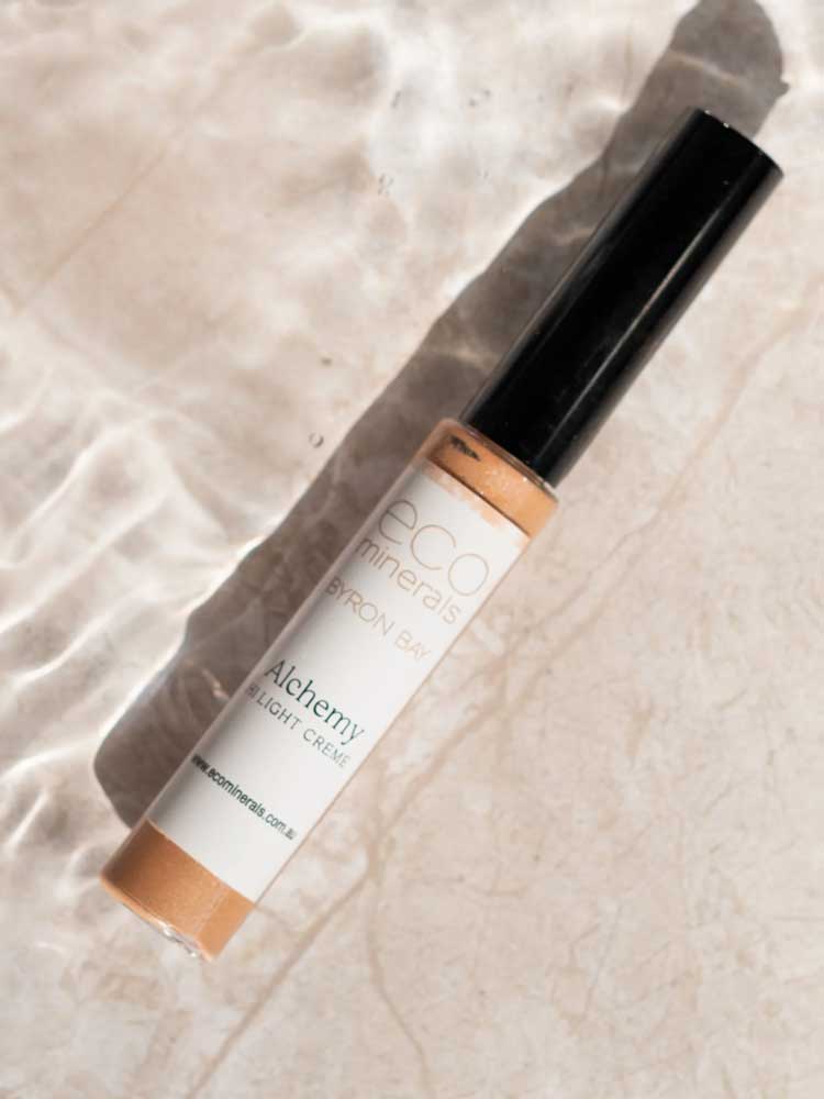 An 8g tube of Eco Minerals Mineral Alchemy High Light Creme, ideal for a radiant finish, rests on a light surface with shimmering water reflections.