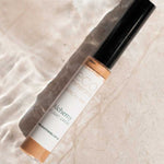 An 8g tube of Eco Minerals Mineral Alchemy High Light Creme, ideal for a radiant finish, rests on a light surface with shimmering water reflections.