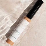 An 8g tube of Eco Minerals Mineral Alchemy High Light Creme, ideal for a radiant finish, rests on a light surface with shimmering water reflections.
