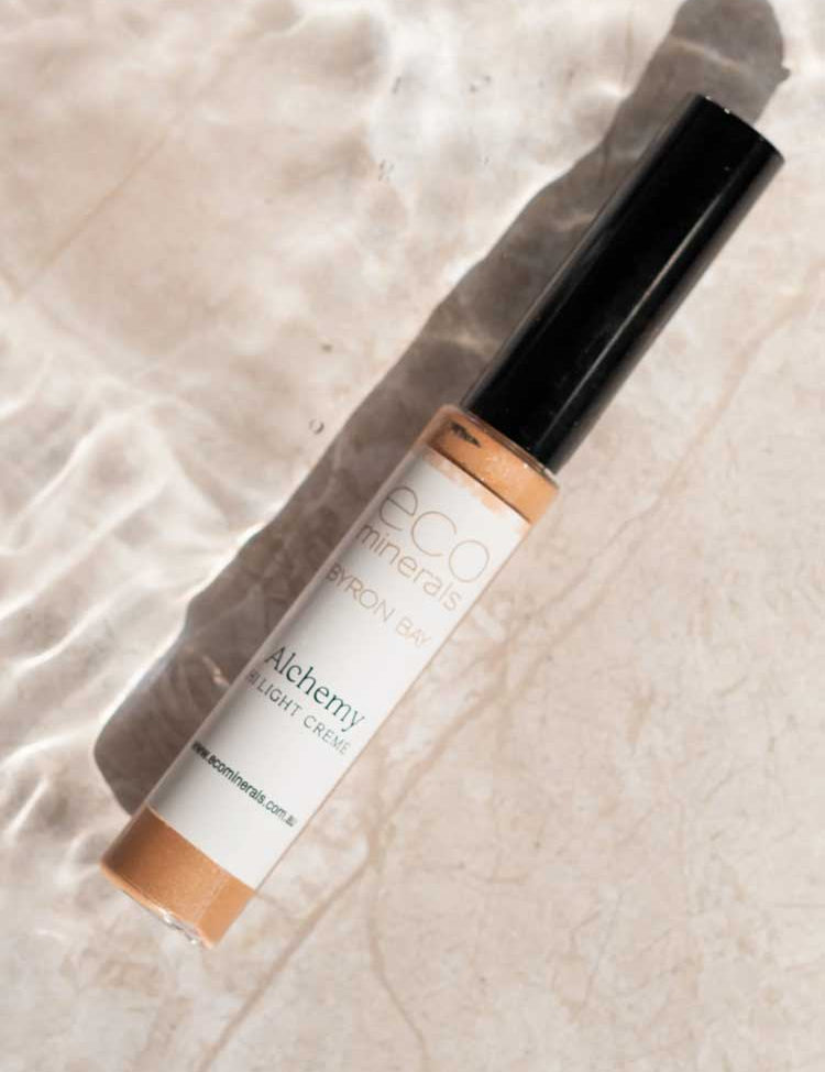 An 8g tube of Eco Minerals Mineral Alchemy High Light Creme, ideal for a radiant finish, rests on a light surface with shimmering water reflections.