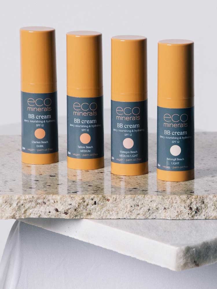 Four bottles of Eco Minerals Mineral BB Cream 32ml in yellow and navy packaging stand upright on a stone surface, each featuring a different shade: Clark Beach, Talow Beach, Wategos Beach, and Belongil Beach.