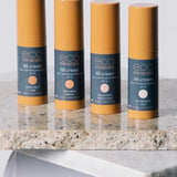 Four bottles of Eco Minerals Mineral BB Cream 32ml in yellow and navy packaging stand upright on a stone surface, each featuring a different shade: Clark Beach, Talow Beach, Wategos Beach, and Belongil Beach.