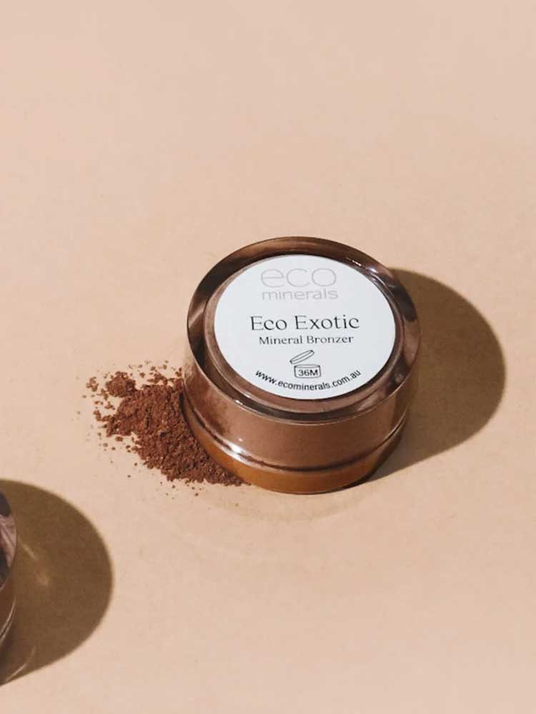 A round container of Eco Minerals Mineral Bronzer - Eco Exotic 4g, a talc-free vegan bronzer, sits on a beige surface with loose bronzer powder spilled beside it.