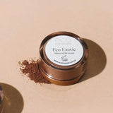 A round container of Eco Minerals Mineral Bronzer - Eco Exotic 4g, a talc-free vegan bronzer, sits on a beige surface with loose bronzer powder spilled beside it.
