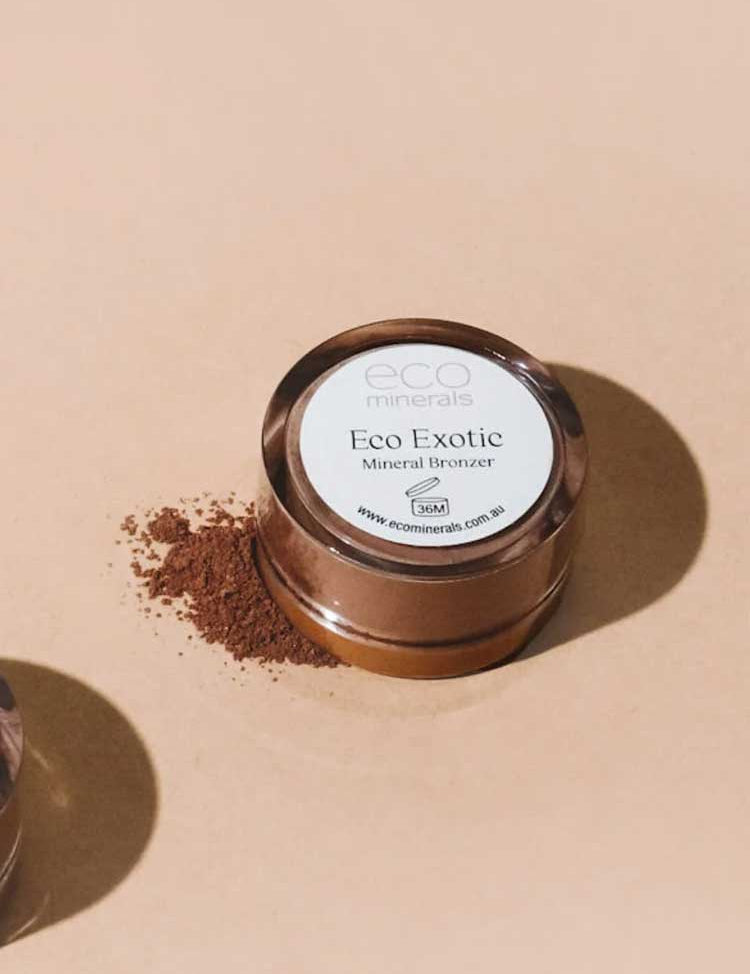 A round container of Eco Minerals Mineral Bronzer - Eco Exotic 4g, a talc-free vegan bronzer, sits on a beige surface with loose bronzer powder spilled beside it.