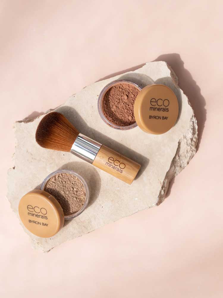 Two containers of Eco Minerals Mineral Bronzer - Eco Exotic 4g and a bamboo makeup brush are arranged on a light stone slab, set against a soft beige background.