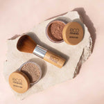 Two containers of Eco Minerals Mineral Bronzer - Eco Exotic 4g and a bamboo makeup brush are arranged on a light stone slab, set against a soft beige background.