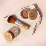 Two containers of Eco Minerals Mineral Bronzer - Eco Exotic 4g and a bamboo makeup brush are arranged on a light stone slab, set against a soft beige background.
