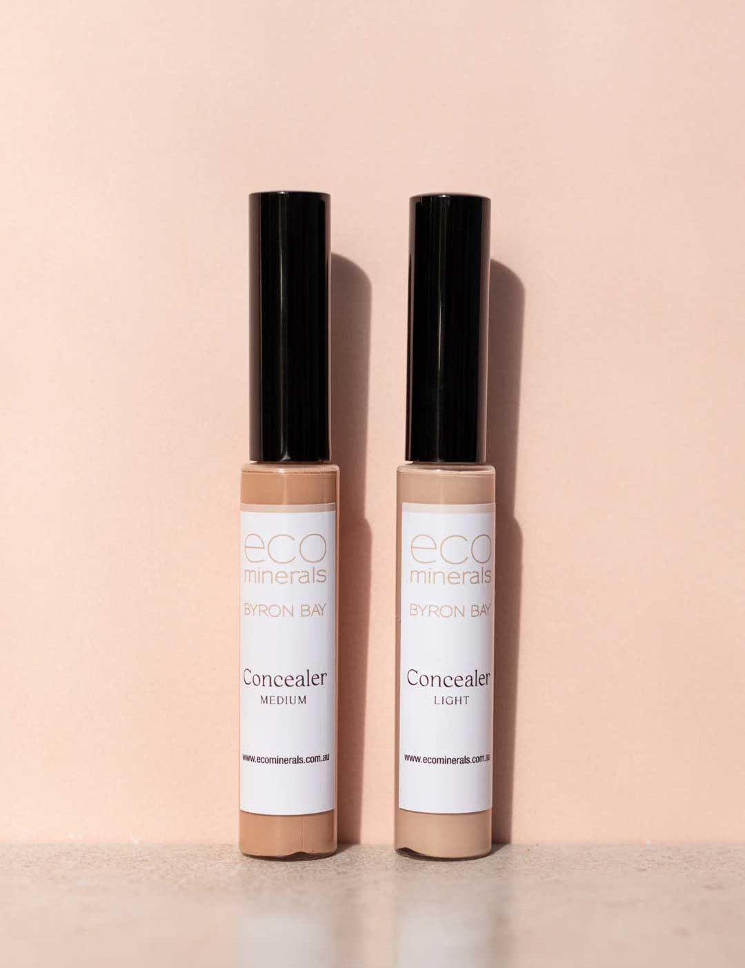 Two tubes of Eco Minerals Mineral Concealer 5g, one in Medium and one in Light, stand upright on a soft peach background with their black caps casting shadows.