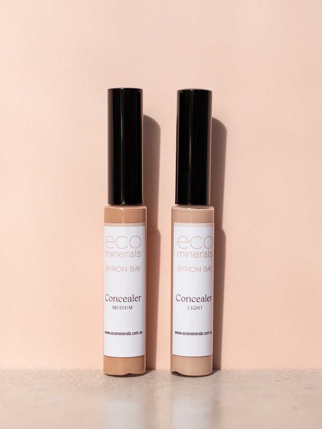 Two tubes of Eco Minerals Mineral Concealer 5g, one in Medium and one in Light, stand upright on a soft peach background with their black caps casting shadows.